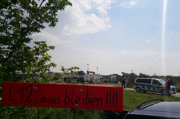 Banner: "L12 muss bleiben", im Hintergrund ein Polizeiwagen beim Materiallager nache dem ehemaligen Dorf Lützerath