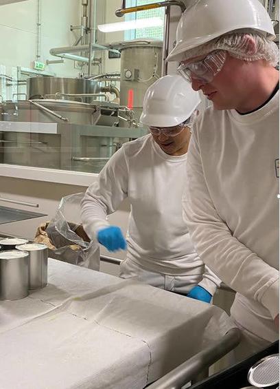 Two men in white outfits, hard hats and safety specs at a canning line with spray driers in the background