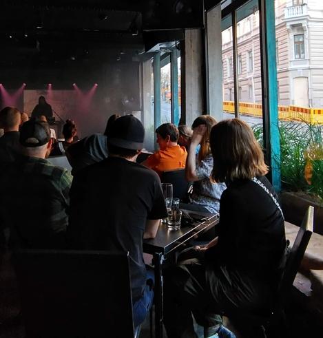 People in dark clothes seated in a small club looking at a man with keyboards and purple lights onstage, to the right a long window with green plants, sun and street signs outside