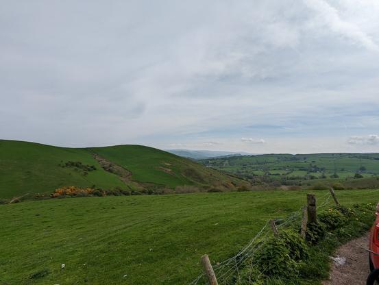Rolling green hills with a wire fence in the foreground and clouds above.