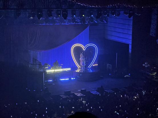 Sabrina Carpenter standing on stage in front of a heart shaped mirror, with yellow LEDs outlining the left side and blue LEDs outlining the right