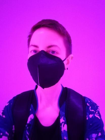 A selfie showing my upper body. I'm a slender white person with short hair wearing a black FFP2 mask and a blue college jacket with a flower pattern. Due to the unique lighting situation at the venue, everything is tinted pink.