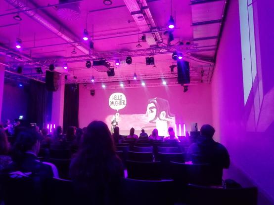 A look at the stage where someone is currently giving a talk. Behind them there's a large projection showing a screenshot of their game: A woman sitting in a bathtub, her head partially submerged in water. In front of her is a rubber duck and over the duck there's a speech bubble that says hello daughter.
In front of the stage, there are rows of chairs, some of them empty.