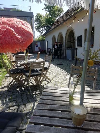 A photo of the outside area of the venue with wooden tables and chairs as well as large parasols. On the table in front of me is a glass with coffee inside it. The sky is clear and the sun shining.