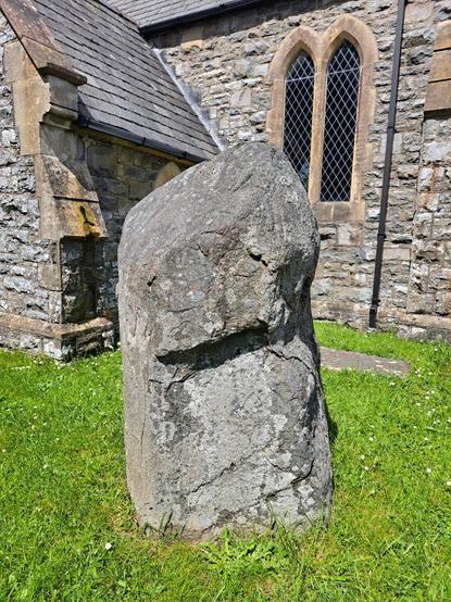 Portrait. A grey stone of close-grained stone standing in a churchyard near the church porch. The stone is a bit less than 6 feet tall, and very broad and chunky. From this angle the top third appears wider than the bottom, coming to a tapered point. The effect is rather phallic.
