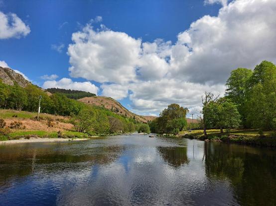 Steep hills drop down to a blue-green river. The water is calm and mirrors the hills and the cloud-strewn blue sky above.