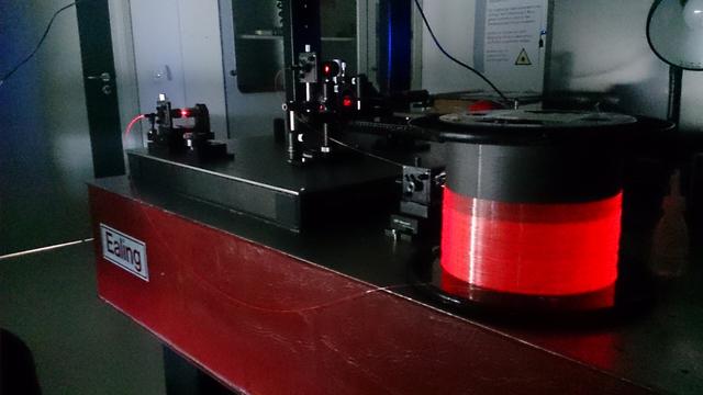 Beschreibung: Ein großer Tisch in einem Labor. Im Vordergrund ist eine rot leuchtende Faser um einen Zylinder gewickelt. Im Hintergrund verlaufen die beiden Enden der Faser zu einem Laser beziehungsweise zu einem Detektor.

Kommentar: Wir haben einen Laserstrahl in eine Glasfaser eingeleitet und gemessen wie viel Licht am Ende noch rauskommt. Aus dem Ergebnis konnten wir die Länge der Faser schätzen.