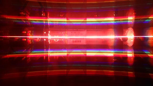 Beschreibung: Im Hintergrund ist ein eine Laserröhre. Im Vordergrund sind verschiedenfarbige Lichtstrahlen.

Kommentar: Das ist der Laser aus meinem Titelbild durch ein optisches Gitter fotografiert. Durch das Gitter haben die verschiedenen Wellenlängen des Lasers ihre Maxima an verschiedenen Stellen. In der Mitte sind alle Wellenlängen des Lasers abgebildet. Dass die Linie rot ist, liegt daran, dass die Wellenlänge 633 nm eine viel größere Leistung hat als die anderen Wellenlängen. Es handelt sich nämlich um einen Helium-Neon-Laser.
