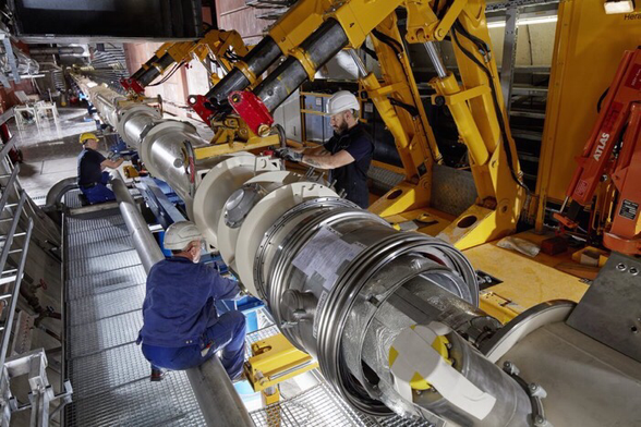 Engineers and technicians install a portion of the beamline for the ALPS experiment in this picture from 2020. This section of the beamline is a long, cylindrical tube that is highly insulated and has an outer shell that looks like a pipe, but is actually a cryogenically cooled magnet setup for the experiment. Three people wearing hard hats guide the installation alongside a support vehicle.