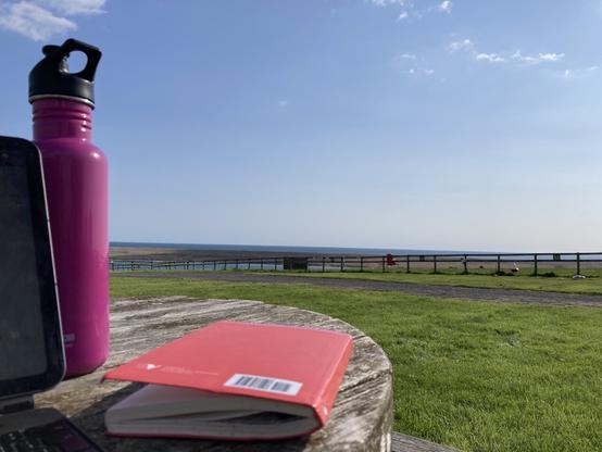 Photo of the view out sea across the green pasture of the campsite and field. On the left is the corner of my iPad, to its right my water bottle, and my notebook sits on the table
