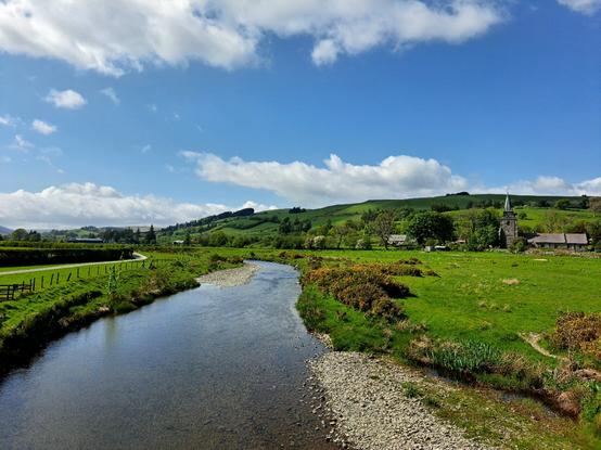 A blue river curves gently away from the viewer up a lush valley. A grey slate church with a tower topped with a short spire is on the right hand side. A blue sky with fluffy white clouds overhead.