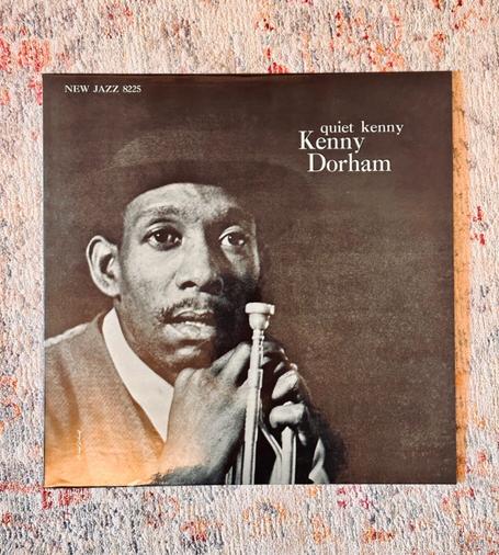 Photo of a record album jacket laying on a pale rug. 

The jacket is a monochrome photo of Kenny Dorham wearing a top hat and holding his trumpet against his face. 

In the top left hand corner, against a dark grey background, white text reads: ‘Quiet Kenny, Kenny Dorham’.
