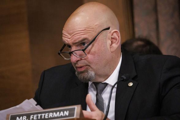 Senator John Fetterman, in a black suit and grey tie, looking sternly over hastily put-on reading glasses at a hapless banker.