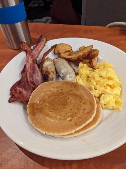 A plate of breakfast food, including pancakes, bacon, eggs, sausage and potato wedges.