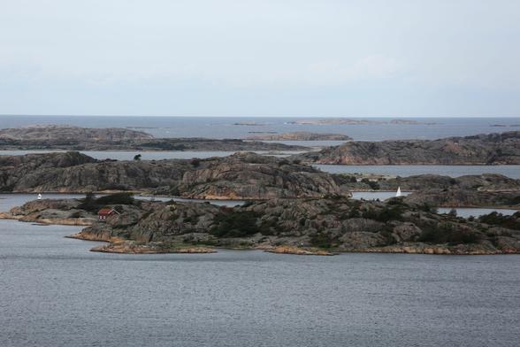 View over a couple of islets which consist mainly of grey round rocks. On the skerry in front a small red cabin is visible. Behind it the top of a sail can be seen indicating a passing sailing boat. 
The water is calm and of a greyish colour.