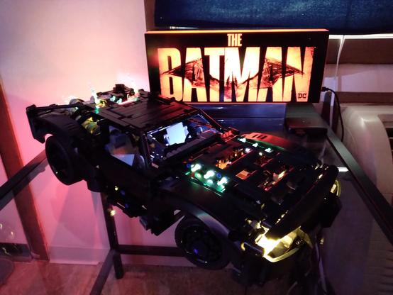 This Lego Technic Batmobile from  the 2022 film "The Batman" is virtually all jet black with the exception of LED lighting that brings out details of the headlights, flametipped exhaust ports, and rear-mounted turbine engine. A lit Batman logo sign from the 2022 film is offset behind and to the right.
