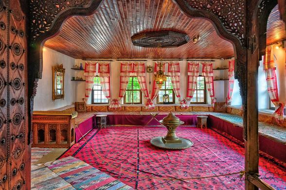 An interior view of a large well lit sitting room with low padded benches along three walls. There are four windows on the far and righthand wall providing natural light. The dominant red color is provided by a large patterned carpet. In the center of the room is a large circular brass incense burner. The scene is framed by an ornately carved entryway.