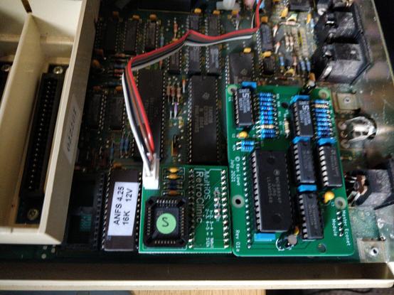 Close up of the inside of an 80s BBC Master 128, fitted with a modern Econet module by Ken Lowe, an ANFS 4.25 ROM installed in slot IC27, as well as a modern quad-boot OS from RetroClinic in the OS ROM socket, with a cable leading to an external switch