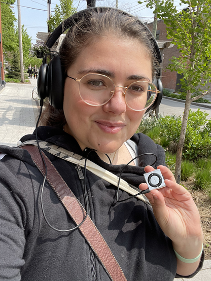Selfie of myself, hair tied back and wearing big headphones, holding up a small silver 2nd Generation iPod Shuffle I bought some time in 2009 and which I am getting back into the habit of using.