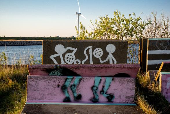 In the foreground, huge stone blocks, used to stop traffic. Now  in storage in the edgelands. The one in the back with stick figures playing tennis. The one in the front has feet, and is placed so that it looks like the feet of the tennis players. In the middle ground sea, and in the background a windmill.
