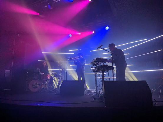 A photo of a musical act on stage in a dark room from maybe 10 feet away. On stage the main act, Jimmy Lavelle, stands leaning, silhouetted behind a mic and keyboards, next to a trumpet player and drum player. Behind them and flanking them are a series of bars of light that look like scratches in the dark through which light is shining through. Fog in the air glows with the pinks and blues of gel stage lights.