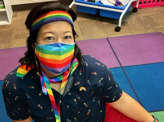 Wisconsin teacher Melissa Tempel says she'll be fired after criticizing her district's decision to block kids from singing "Rainbowland." Melissa Tempel is wearing a t-shirt with mini rainbow icons, a rainbow pride face mask, headband, ascot and lanyard with smiling eyes.