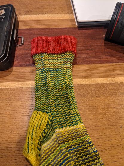 Folded over red cuff on a hand knit sock that otherwise is yellow and browns and blues sitting on a wooden table.