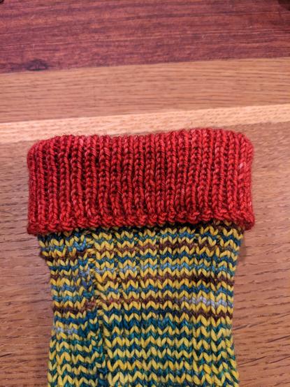 Folded over red cuff on a hand knit sock that otherwise is yellow and browns and blues sitting on a wooden table. A close up of just the cuff in this shot.