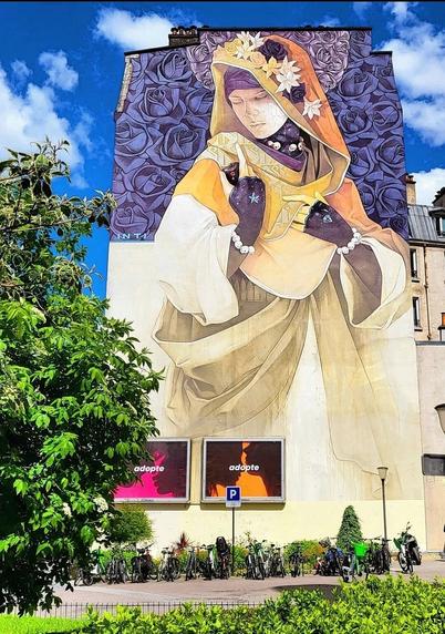 Streetartwall. On the outer wall of a narrow multi-storey house is sprayed/painted a mural of an elegant lady.  The upper part is a frieze of purple roses, below a light yellow background. Borrowing from Art Nouveau, on it is a lady in a lush dress in light yellow and striking drapery and wide sleeves. The dress merges into a bright orange cloth with bright flowers on it, which then covers her head. Her hair is under a purple colored cloth and her bright, finely drawn face is encompassed by a scarf in purple underneath, on which tiny white skulls can be seen. Her hands reach into the cloth of her dress in front of her chest. She wears Iila colored gloves. Here, too, the beaded bracelets on them can be recognized as skulls on closer inspection. A star on one hand and a diamond on the other complete the whole.
A very elaborate and detailed Mural. Title: "La Madre secular 2"