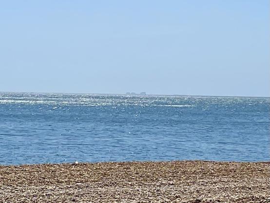 Blue sky, sunshine glittering on the little waves of a various blue sea. Pebbles of the beach in the foreground. In the distance is the shadowy shape of Dungeness Power Station.
