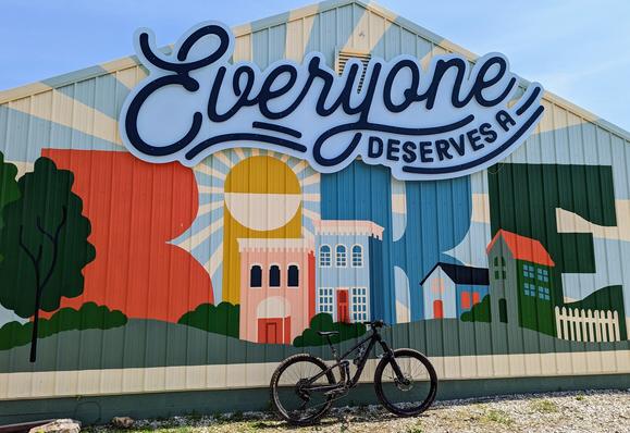 Mountain bike leaning on the side of a building with a mural. At the top it says "Everyone deserves a", The mural is a small community of buildings stylized to spell the word "BIKE"