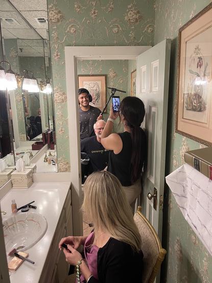 Photo of a people in a powder room and a bathroom. A man wearing headphones and holding a shotgun mic on a boom is in the bathroom. A woman facing away is holding a mobile phone, taking a photo. A another woman in the foreground sits at the powder room sink and faces away, at the others.