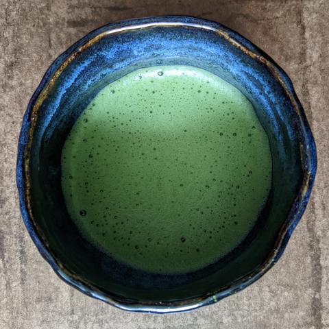 Overhead shot of a tea bowl with dark blue glaze that has oxidized red-gold and pale blue rings. Its rim is round but has a clearly hand-made wobbling/wavy shape to it. Inside the bowl is a frothy light green matcha. A bit of a shadow falls over the bowl.