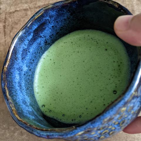 Closeup of a hand tilting the teabowl to show off the soft yet vibrant colour of the frothy light green matcha against the dark blue of the bowl.