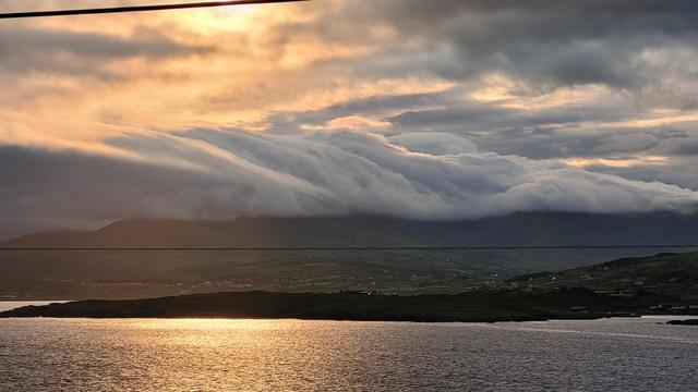 Clouds like a waterfall over the hills. Above orange light from the sunrise. The wster of the bay has a an orange glow
