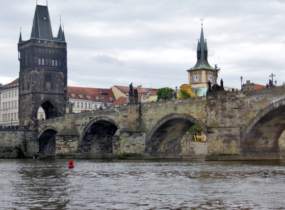 A Stone arch medieval bridge on river Vltava. On the left side at the entrance of bridge has a covered tall structure. Behind the river some buildings are seen.