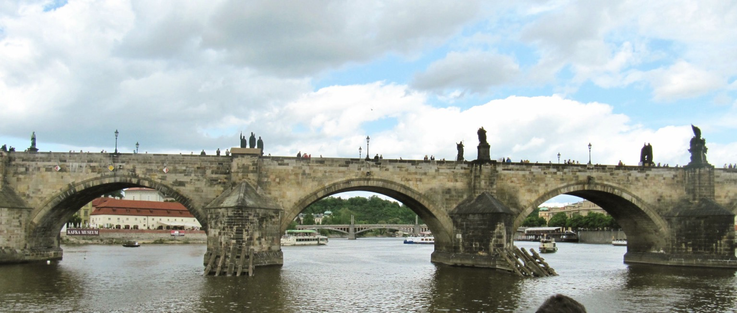 Charles bridge, boats are seen on the river   Statues on the bridge, Blue and white clouds above.