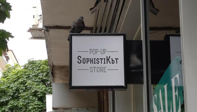 A city dwelling pigeon perched on a boutique's street sign inscribed "Pop-Up SophistiКът Store."