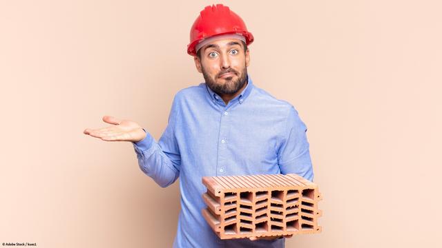 Ein Mann in einem blauen Hemd und mit einem roten Bauhelm auf dem Kopf. Er hält einen Ziegelstein in der linken Hand und schaut mit fragendem Blick in die Kamera.