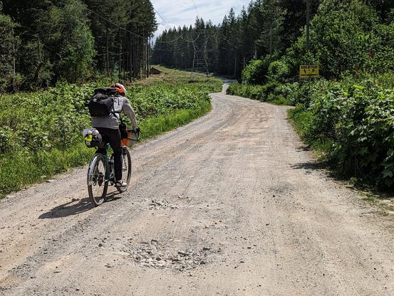 a wide gravel road beneath powerlines that goes through a forest; a cyclist on a single-speed gravel bike carries on