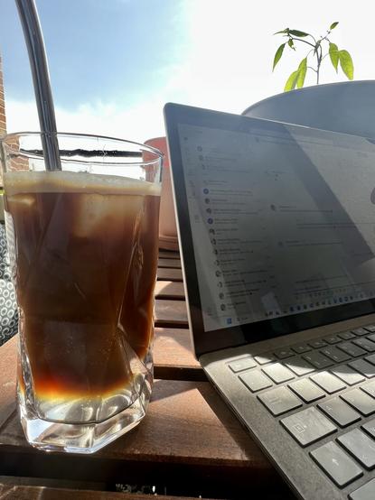 Nahaufnahme von einem Glas Espresso-Tonic mit einem Metallsteohhalm neben einem Laptop auf einem Holztisch. Im Hintergrund blauer Himmel und eine Chili-Pflanze