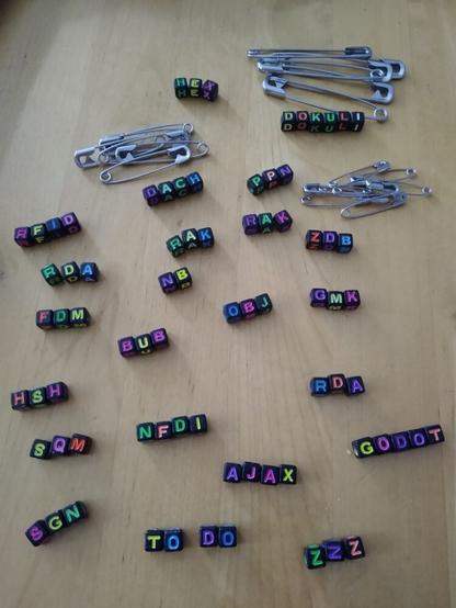 Schwarze quadratische Perlen mit neonfarbenen Buchstaben darauf und eine Handvoll Sicherheitsnadeln auf einem Holztisch.