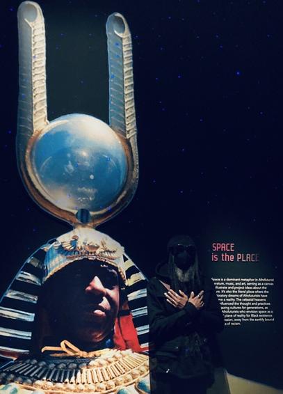 A photo of myself standing in front of a large image of Sun Ra with Egyptian head piece featuring an iridescent globe and two pronged fork or antenna.