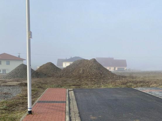 Das Ende einer asphaltierten Straße, an deren linker Seite ein rot gepflasterter Gehweg verläuft. Links ein Mast. Im Hintergrund, etwas im Nebel liegend, drei Einfamilienhäuser. In der Bildmitte drei Hügel mit Erdaushub.