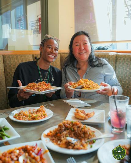 Yvonne Turner and owner of Uptown China posing with plates of fried rice and noodles in a booth, with more plates of food in front of them
