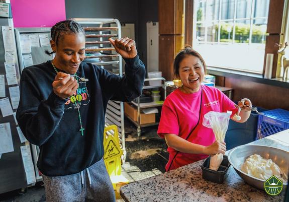 Yvonne Turner (left) holding a spoon and dancing while owner of Fuji Bakery (right) fills a piping bag with white frosting