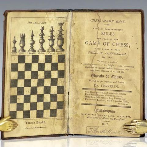 an aging leather bound book. one open page shows the board and pieces, the other shows and illustrative cover page.