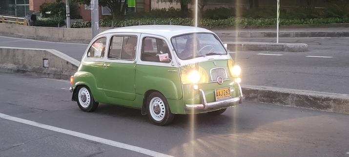 A two-tone, green lower, white upper Fiat 600 Multipla from the 60s, in traffic, with headlights on in late afternoon, looking great!