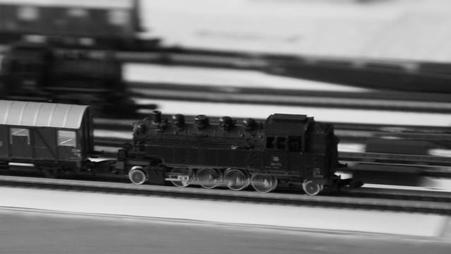 SW - Spielzeuglok in voller Fahrt. Im Hintergrund sieht man verschwommen eine Lok und einen gedeckten Güterwagen