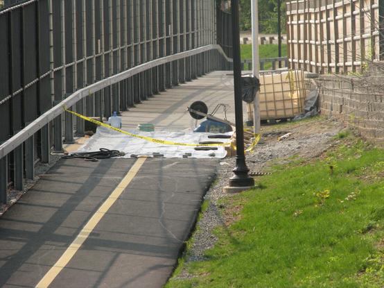 Photo from Lowell St region of Community Path looking east, showing Lowell to Central segment. Where the asphalt path turns to concrete there is plastic sheeting across the path, held down by random objects including hoses and an upside down wheelbarrow. There is caution tape across the path and a large cubical tank of water off to the side.